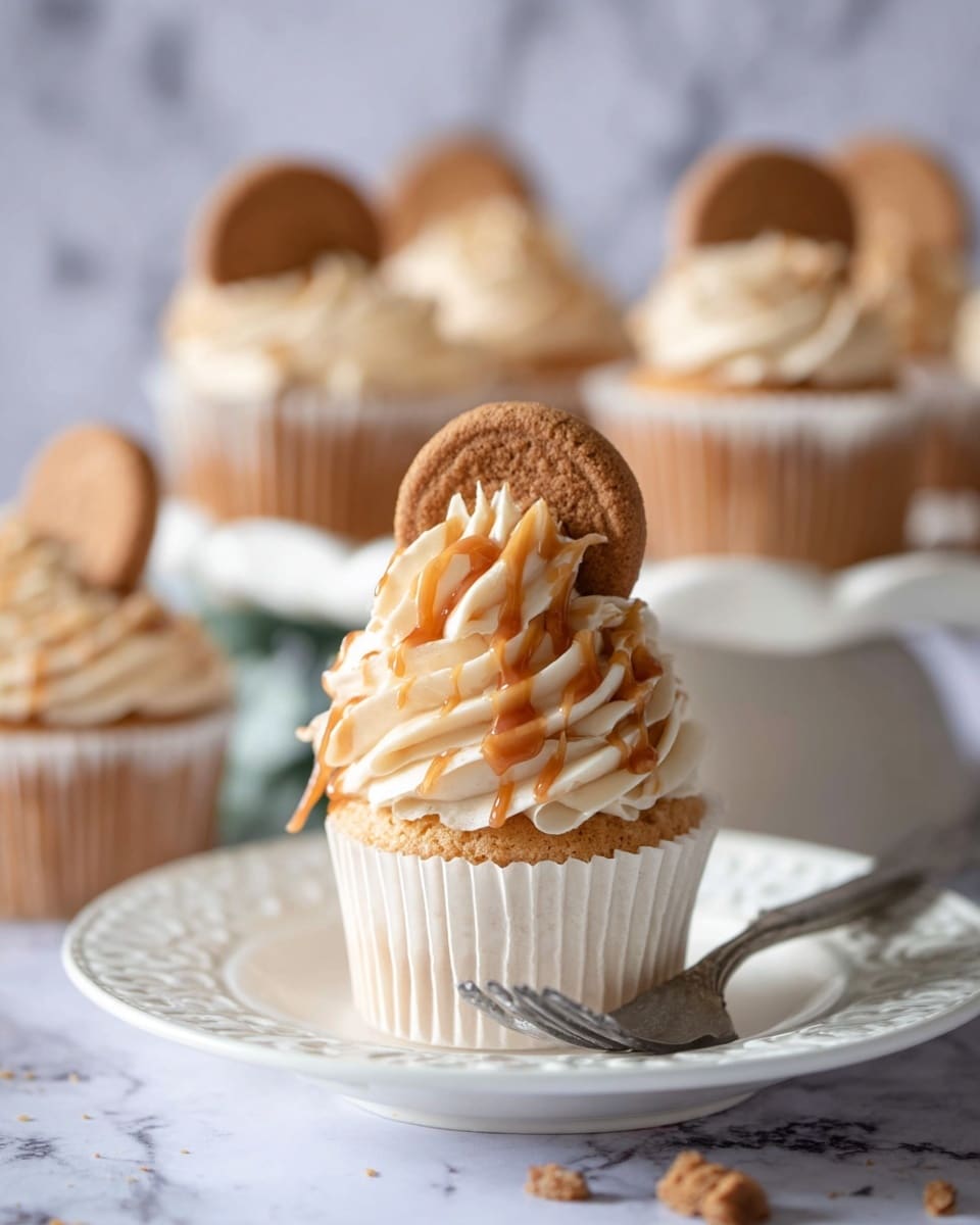 A single cupcake is shown in close-up, sitting on a white decorative plate with a silver fork next to it. The cupcake has a soft, light beige base wrapped in a white paper liner. On top, there is a swirl of creamy, pale beige frosting with a smooth texture. Drizzled over the frosting is a glossy caramel sauce that runs slightly down the sides. A round brown cookie is placed upright in the frosting, adding a crunchy texture to the soft cupcake. In the background, several more cupcakes with the same design are slightly out of focus on a white cake stand. The scene is set on a surface with a white marbled texture. Photo taken with an iphone --ar 4:5 --v 7