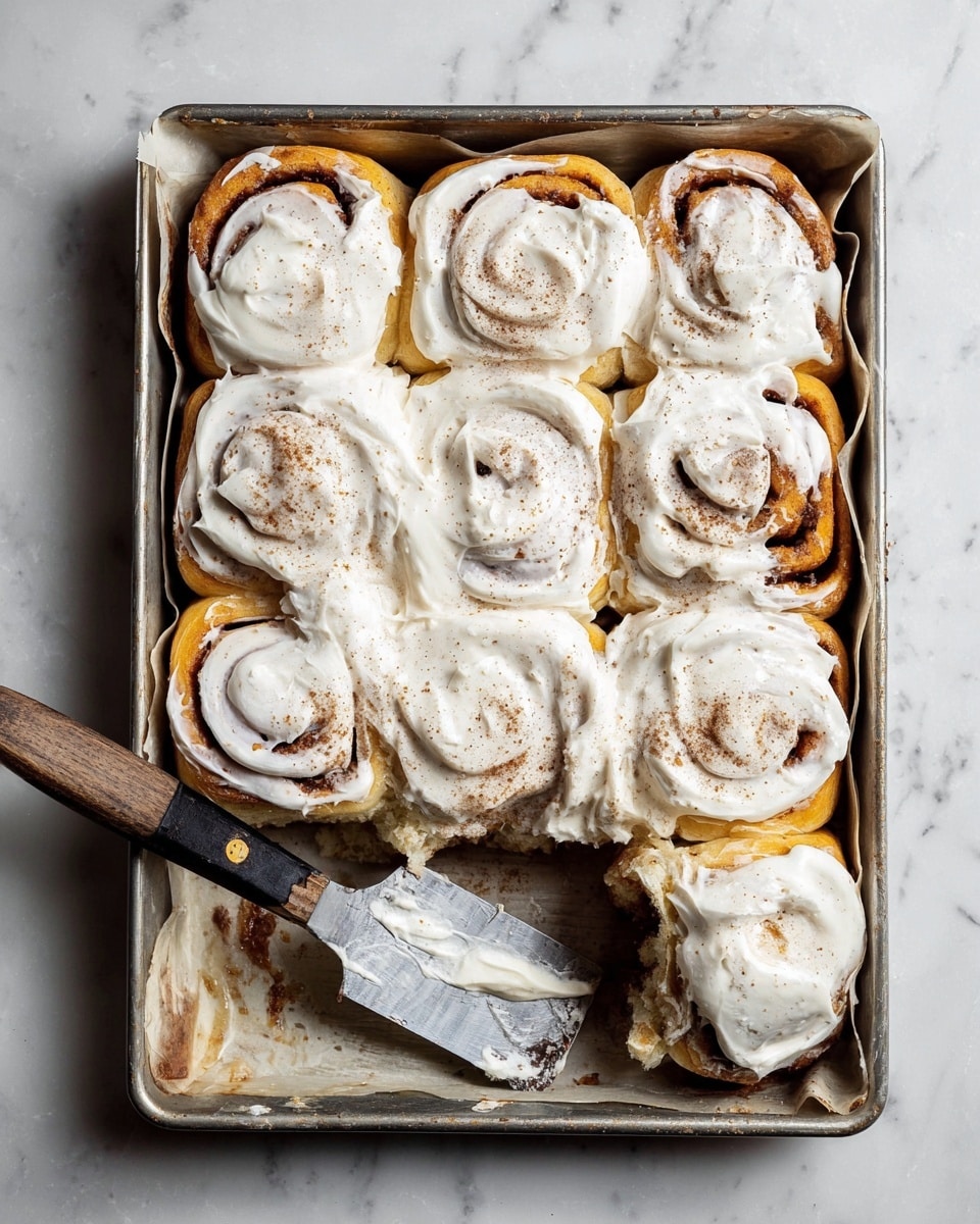 A metal baking pan lined with parchment paper holds nine cinnamon rolls arranged in a 3x3 grid, each roll showing visible layers of golden-brown dough swirled with darker cinnamon filling. A thick white cream cheese frosting generously covers all the rolls, with some spots lightly sprinkled with cinnamon powder. One cinnamon roll in the bottom right corner is slightly pulled out, revealing its soft, fluffy layers more clearly. A metal spatula with a wooden handle lies inside the pan near the bottom left corner, partially smeared with frosting. The pan is placed on a white marbled surface. photo taken with an iphone --ar 4:5 --v 7