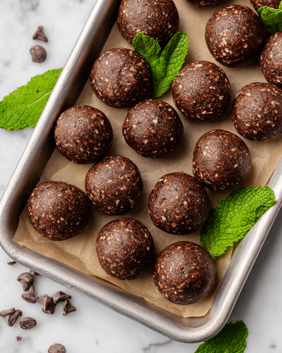 The image shows a metal tray lined with parchment paper holding twelve round, smooth energy balls. Each ball is dark brown with visible small light oat pieces and chocolate chips inside, giving a rough, textured appearance. The balls are arranged in four rows, and several fresh green mint leaves are placed as decoration on and around the tray. The tray is set on a white marbled surface with a few scattered chocolate chips nearby. photo taken with an iphone --ar 4:5 --v 7