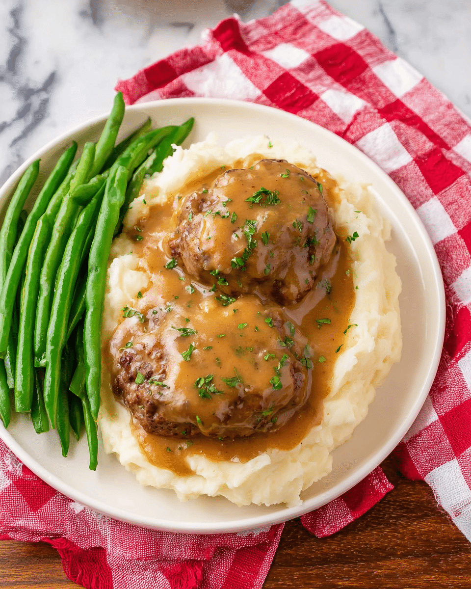 A white plate holds a meal with three main layers: on the bottom, there is a thick, creamy layer of smooth white mashed potatoes spread across the plate; above it, two large, browned meat patties are placed side by side, partially covered by a rich, light brown gravy that has a slightly glossy texture; the gravy is sprinkled with small green herb pieces adding a fresh look; on the left side of the plate, a bunch of bright green steamed green beans with a slight shine complete the dish, all resting on a white marbled surface with a red and white checkered cloth partially under the plate; photo taken with an iphone --ar 4:5 --v 7