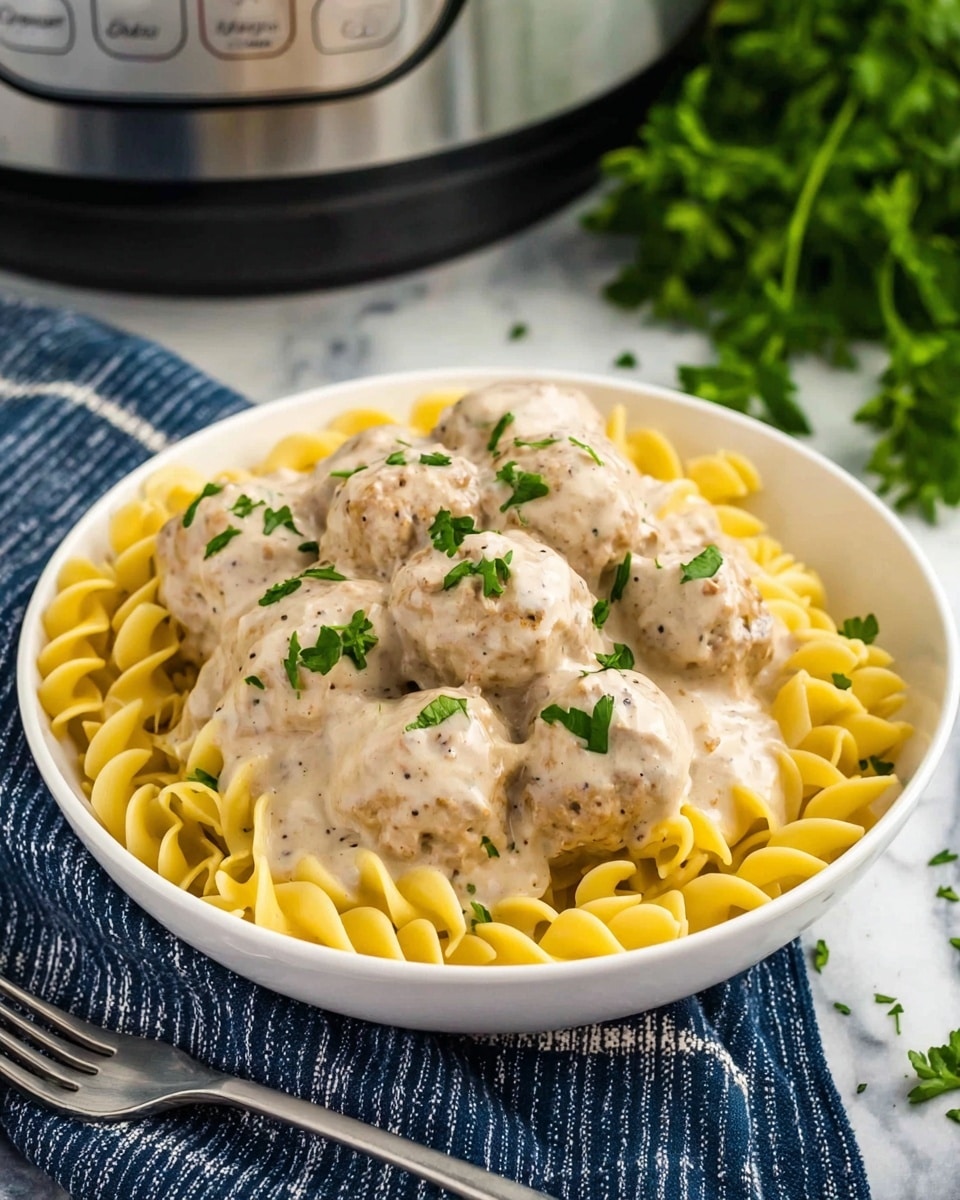 A white bowl filled with a base layer of yellow curly egg noodles with a soft texture, topped by a generous layer of light brown meatballs covered in creamy, beige sauce speckled with black pepper and garnished with small pieces of fresh green parsley. The bowl is placed on a white marbled surface with a fork lying next to it and some green herbs blurred in the background, along with part of a slow cooker and a dark blue cloth with white stripes visible. photo taken with an iphone --ar 4:5 --v 7