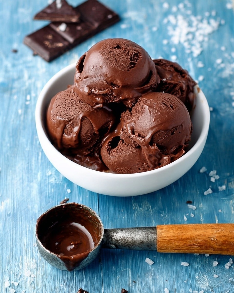 The image shows a white bowl filled with four scoops of rich, dark brown chocolate ice cream, each scoop slightly shiny with a smooth, creamy texture and a few melted spots on top. Next to the bowl is a vintage ice cream scoop with some melted chocolate inside its round metal scoop and a wooden handle. In the upper part of the image, there are a few pieces of broken dark chocolate placed on a white marbled surface that looks like a wood texture painted blue with some white salt flakes scattered around. The photo taken with an iphone --ar 4:5 --v 7