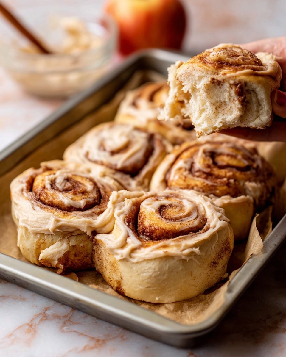 The image shows a tray of six cinnamon rolls with three visible layers: the soft, light beige dough base with a slightly flaky texture, a rich cinnamon-brown spiral layer in the middle, and a creamy light tan frosting spread unevenly on top and around the sides. One cinnamon roll is being lifted by a woman's hand, revealing the swirled dough and cinnamon filling inside. The rolls sit on parchment paper inside a metal tray, placed on a white marbled surface. In the blurred background, there is a glass bowl with a spoon and a whole apple. Photo taken with an iphone --ar 4:5 --v 7