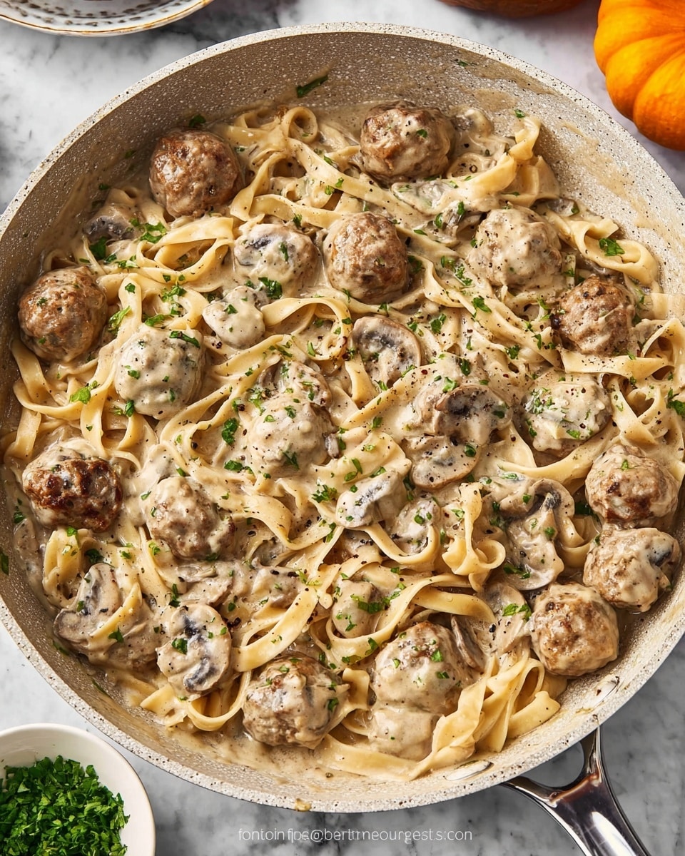 A large frying pan contains a creamy mushroom sauce with many brown meatballs and wide, flat noodles mixed throughout. The sauce is light beige, coating the meatballs, noodles, and sliced mushrooms evenly. Small bits of green herbs are sprinkled on top for color, and black pepper is visible as tiny dark specks. The pan has a light speckled interior and a metallic handle. The background is a white marbled texture with part of an orange pumpkin visible at the top right edge and a small white bowl of chopped green herbs in the bottom left corner. Photo taken with an iphone --ar 4:5 --v 7