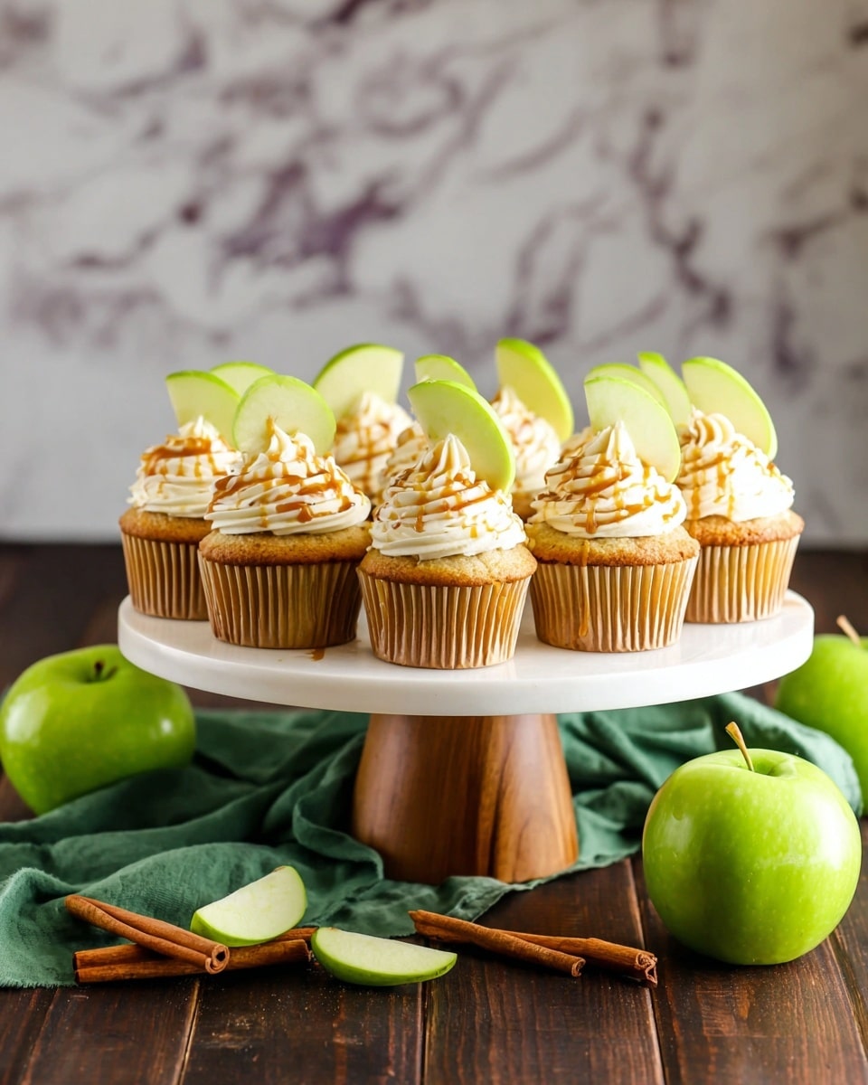 A white cake stand with a wooden base holds nine cupcakes arranged in three rows. Each cupcake has a golden brown base with a swirl of creamy white frosting on top, drizzled lightly with caramel sauce. A thin, bright green apple slice is placed upright into the frosting on each cupcake. Surrounding the stand are whole and sliced green apples and two cinnamon sticks on a folded green cloth, set on a dark wooden surface with a white marbled texture background. photo taken with an iphone --ar 4:5 --v 7