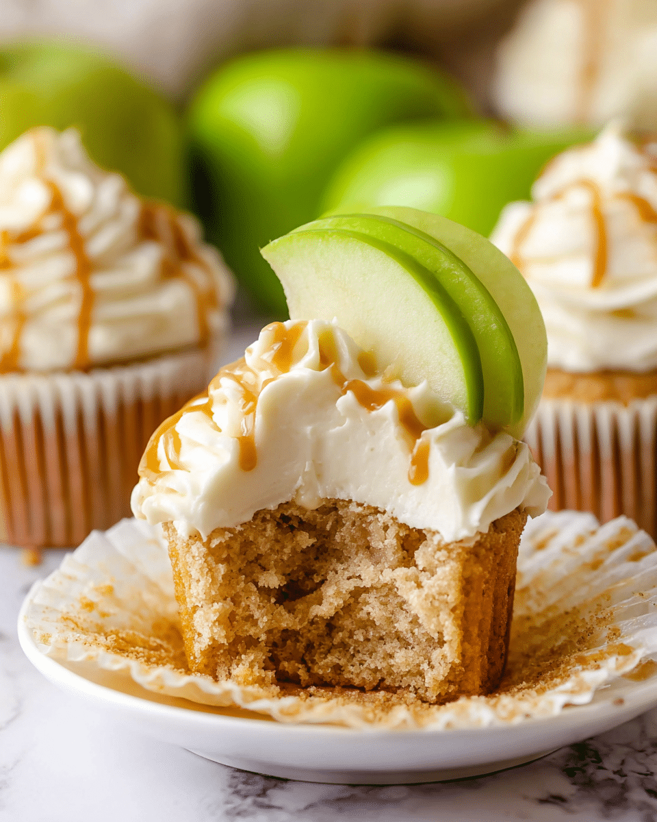 A close-up of a cupcake with one bite taken out shows three main layers: a bottom layer of soft, light brown cake with a slightly crumbly texture, a thick, smooth white frosting layer on top, and thin swirls of caramel sauce drizzled over the frosting. A green apple slice is placed at the back of the frosting, standing upright. The cupcake sits on a white plate with the cupcake liner peeled down around it, all set on a white marbled surface with other whole cupcakes and green apples blurred in the background. photo taken with an iphone --ar 4:5 --v 7