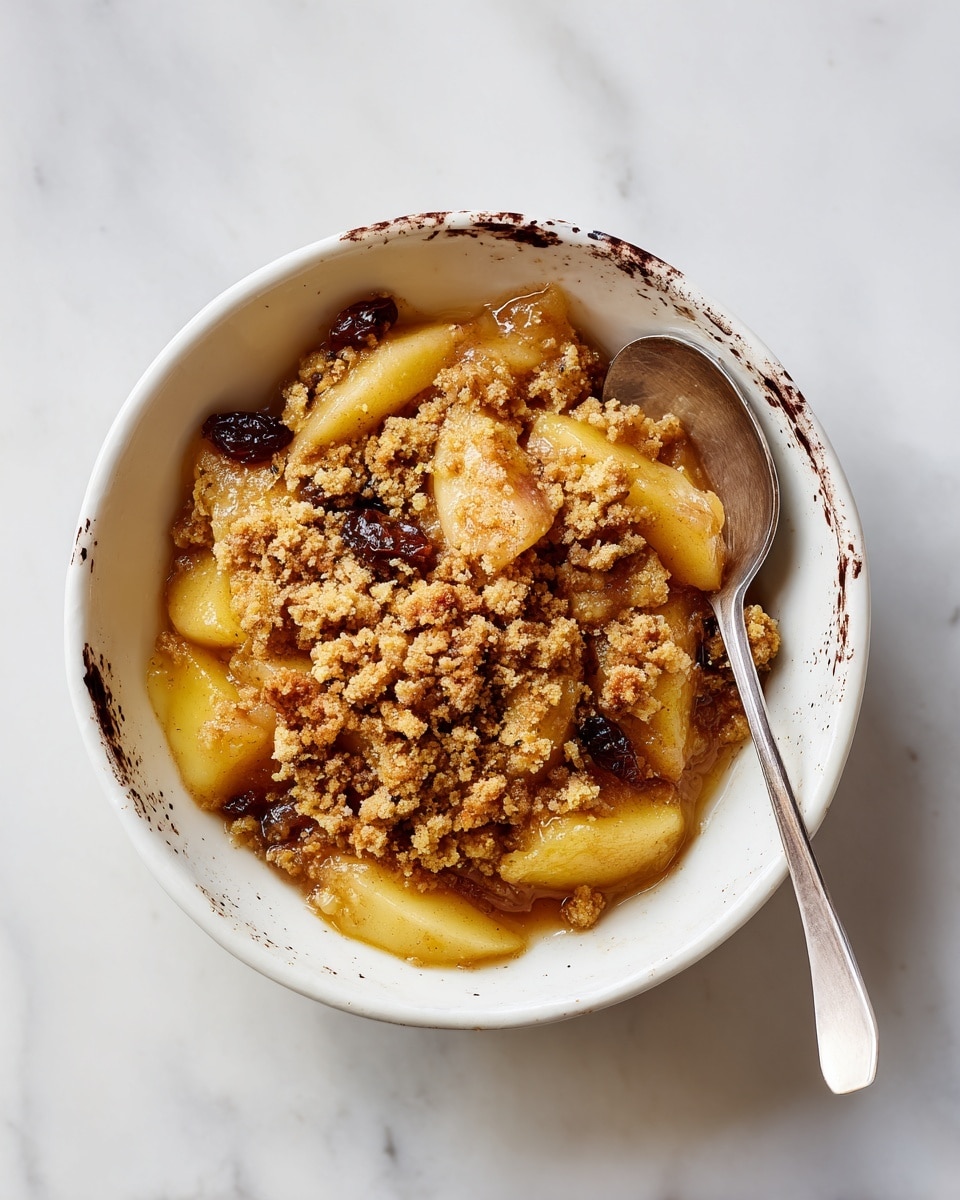 A white bowl with dark brown splashes holds a dish with three main layers: at the bottom, soft cooked apple slices in a caramel-like brown sauce; in the middle, a crumbly, light golden crumble topping; and scattered dark raisins mixed throughout. The textures vary from soft and juicy to crumbly and dry. A silver spoon rests inside the bowl against the right edge. The bowl is placed on a white marbled surface. Photo taken with an iphone --ar 4:5 --v 7