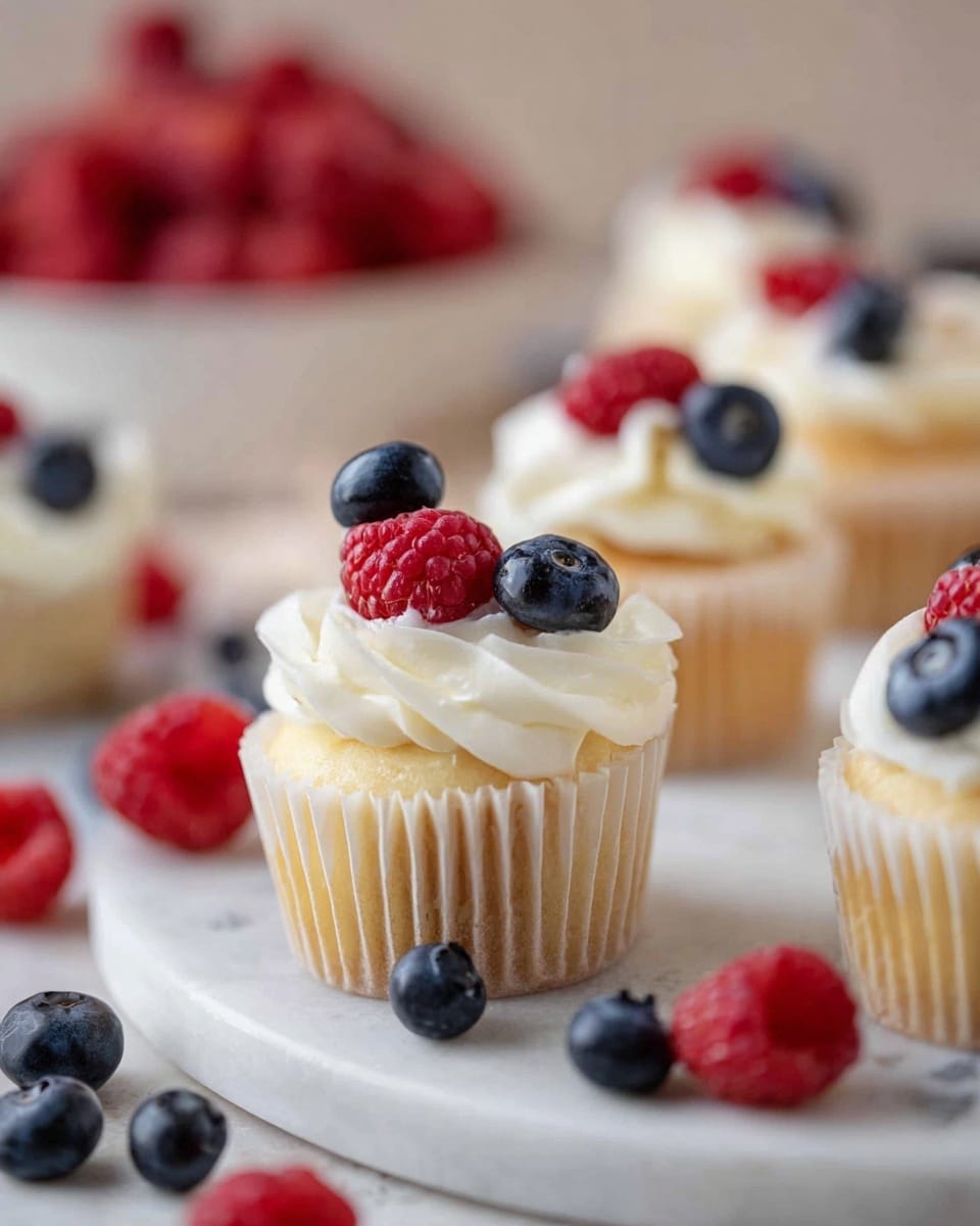 Several small vanilla cupcakes sit on a round white marble surface, each with a light golden base wrapped in white paper liners. Each cupcake is topped with a thick swirl of smooth white frosting, layered with a fresh raspberry and a halved blueberry placed carefully on top. Around the cupcakes, loose blueberries and raspberries add pops of deep blue and bright red color, creating a fresh and colorful look. In the background, a white bowl filled with more raspberries is slightly blurred, emphasizing the main small cupcakes in the front. Photo taken with an iphone --ar 4:5 --v 7