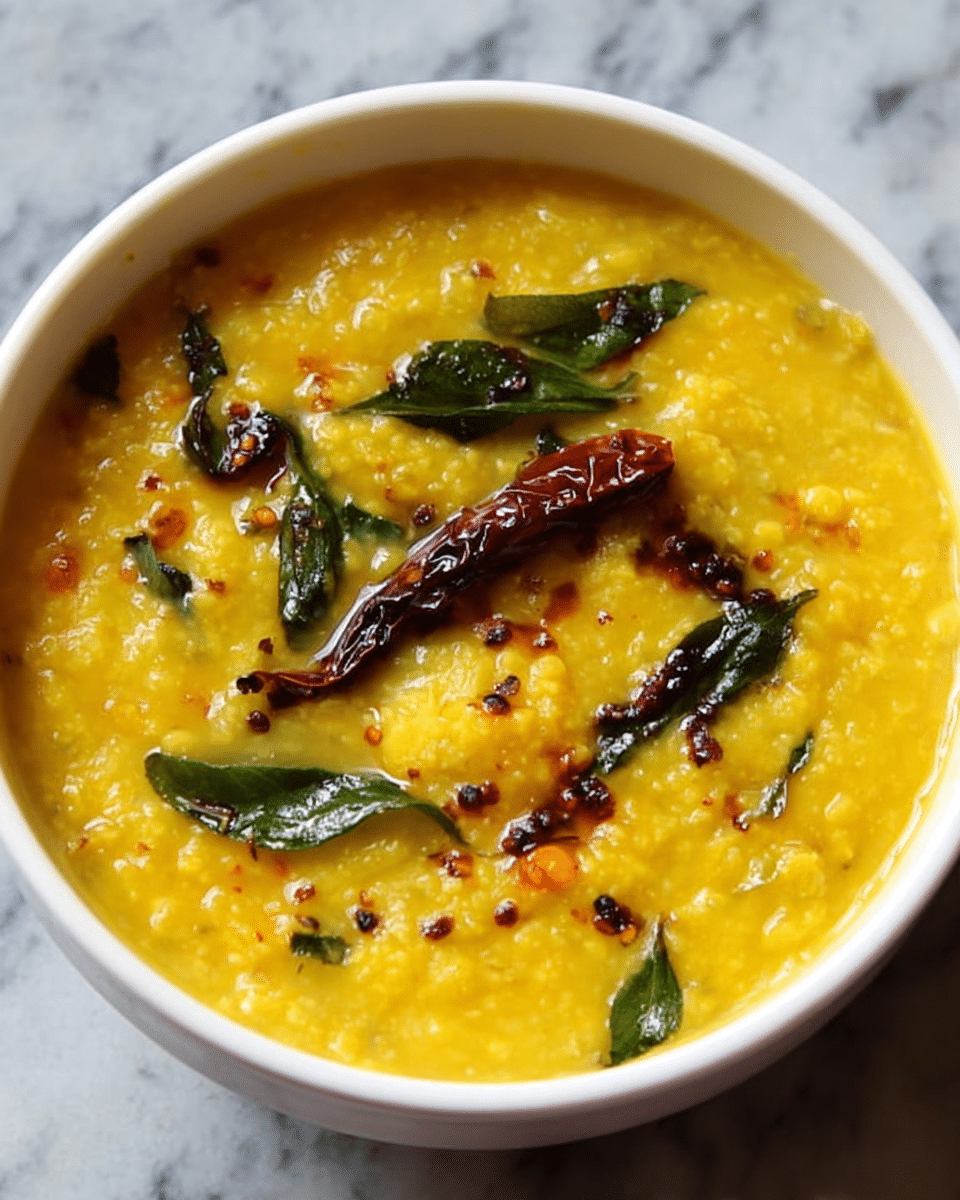A white bowl filled with thick yellow lentil soup showing small red dried chilies and mustard seeds floating on top, along with some green curry leaves. The texture is creamy with some visible mustard seeds and lentils, and the soup looks warm and rich. The bowl is placed on a white marbled surface. Photo taken with an iphone --ar 4:5 --v 7