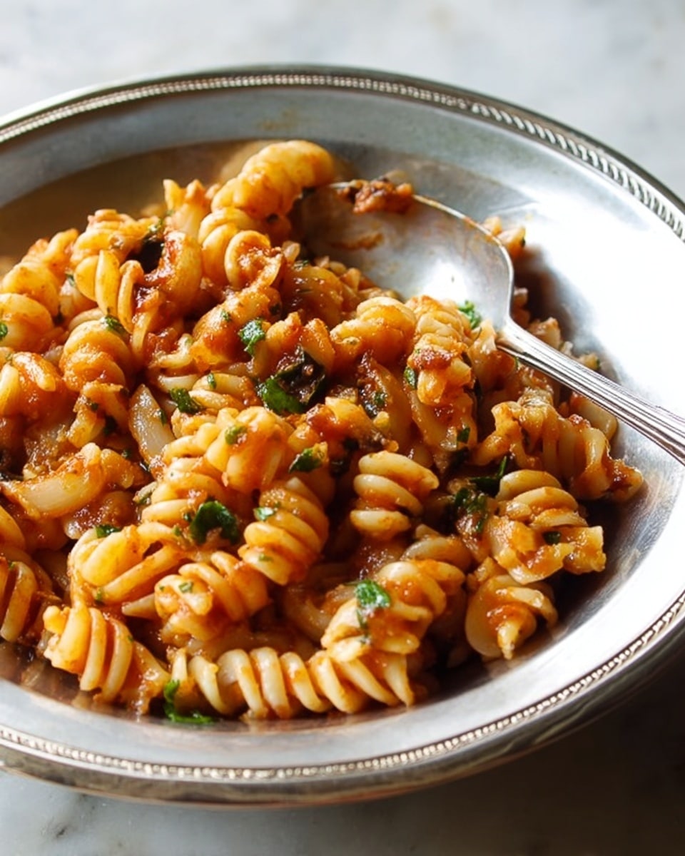 A close-up view of spiral pasta coated in a bright red tomato sauce with visible small bits of greens and orange vegetables mixed throughout. The pasta is packed tightly in a shiny silver bowl with dented metal texture showing some reflections. A silver fork rests on the edge inside the bowl, positioned among the pasta. The image background is changed to a white marbled texture. photo taken with an iphone --ar 4:5 --v 7