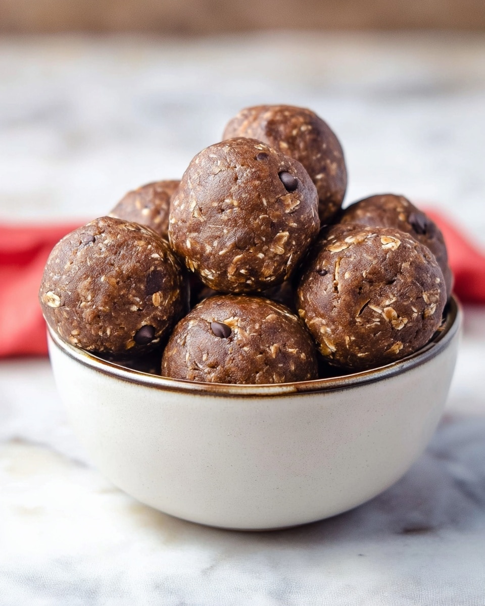 A white round bowl filled with seven chocolate oat balls, each ball showing a rough texture with visible oatmeal pieces and small chocolate chips. The balls are stacked closely inside the bowl, with one ball prominently in the center, slightly larger and positioned higher than the others. The chocolate oat balls have a matte, slightly crackled surface in dark brown color. The bowl is placed on a white marbled surface with soft natural light highlighting the details. Photo taken with an iphone --ar 4:5 --v 7