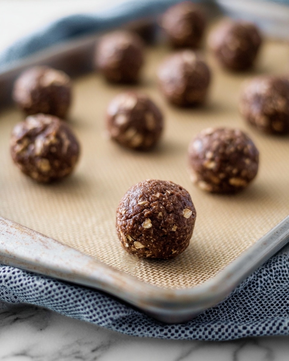The image shows several small, round cookie dough balls arranged on a baking sheet lined with a beige silicone mat. Each ball is chocolate brown with visible oatmeal flakes embedded throughout, giving a rough texture. The baking sheet is metal with a slightly worn edge, sitting on top of a dotted cloth that is draped over a white marbled surface. The photo focuses on the front cookie dough ball, making the ones in the back softly blurred. photo taken with an iphone --ar 4:5 --v 7