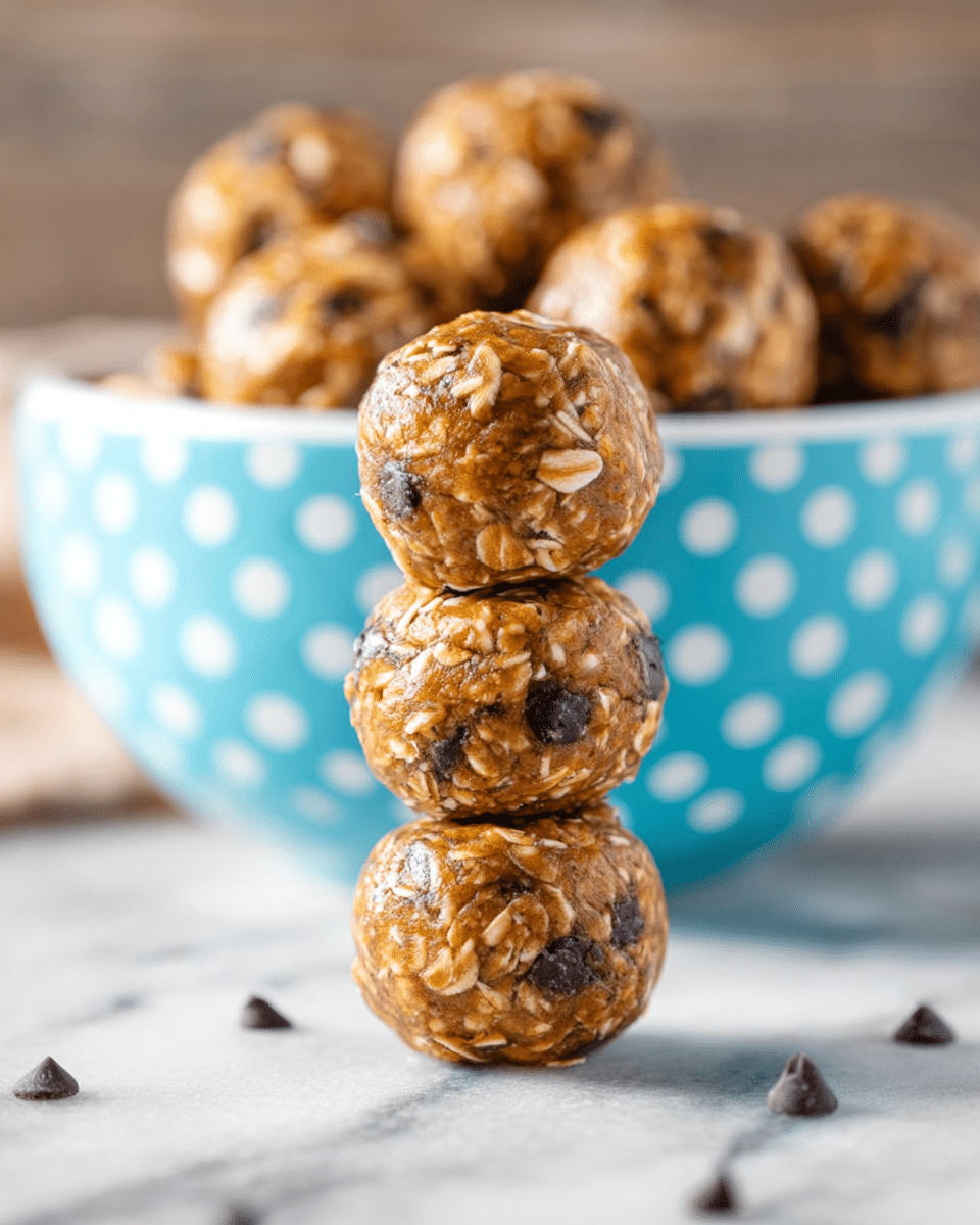 The image shows a small stack of four round energy bites in the front, each with a golden-brown color mixed with visible oats and dark chocolate chips scattered around the surface, giving a textured and slightly glossy look. Behind the stack, a white bowl with light blue and white polka dots is full of more energy bites, all similar in color and texture. The setup is on a white marbled surface, creating a light background that contrasts with the warm tones of the energy bites. photo taken with an iphone --ar 4:5 --v 7