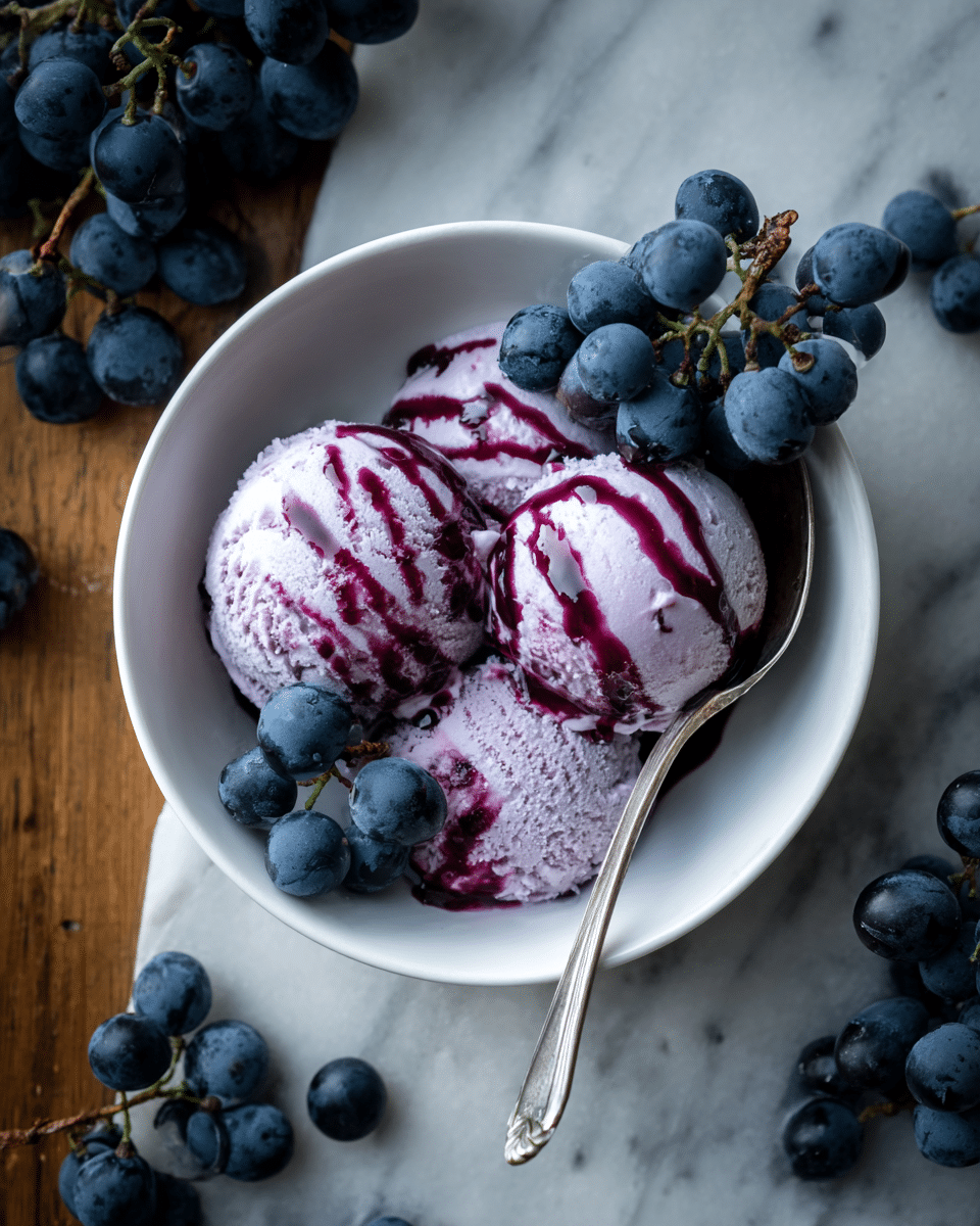 A bowl filled with three scoops of purple ice cream, each topped with dark red syrup drizzled over them, is placed on a white marbled texture. Around and on top of the ice cream are fresh dark blue grapes, adding a contrast of color and texture. A silver spoon rests inside the bowl on the left side. In the background, there is a small jar filled with dark red liquid and some additional grapes scattered softly. Photo taken with an iphone --ar 4:5 --v 7