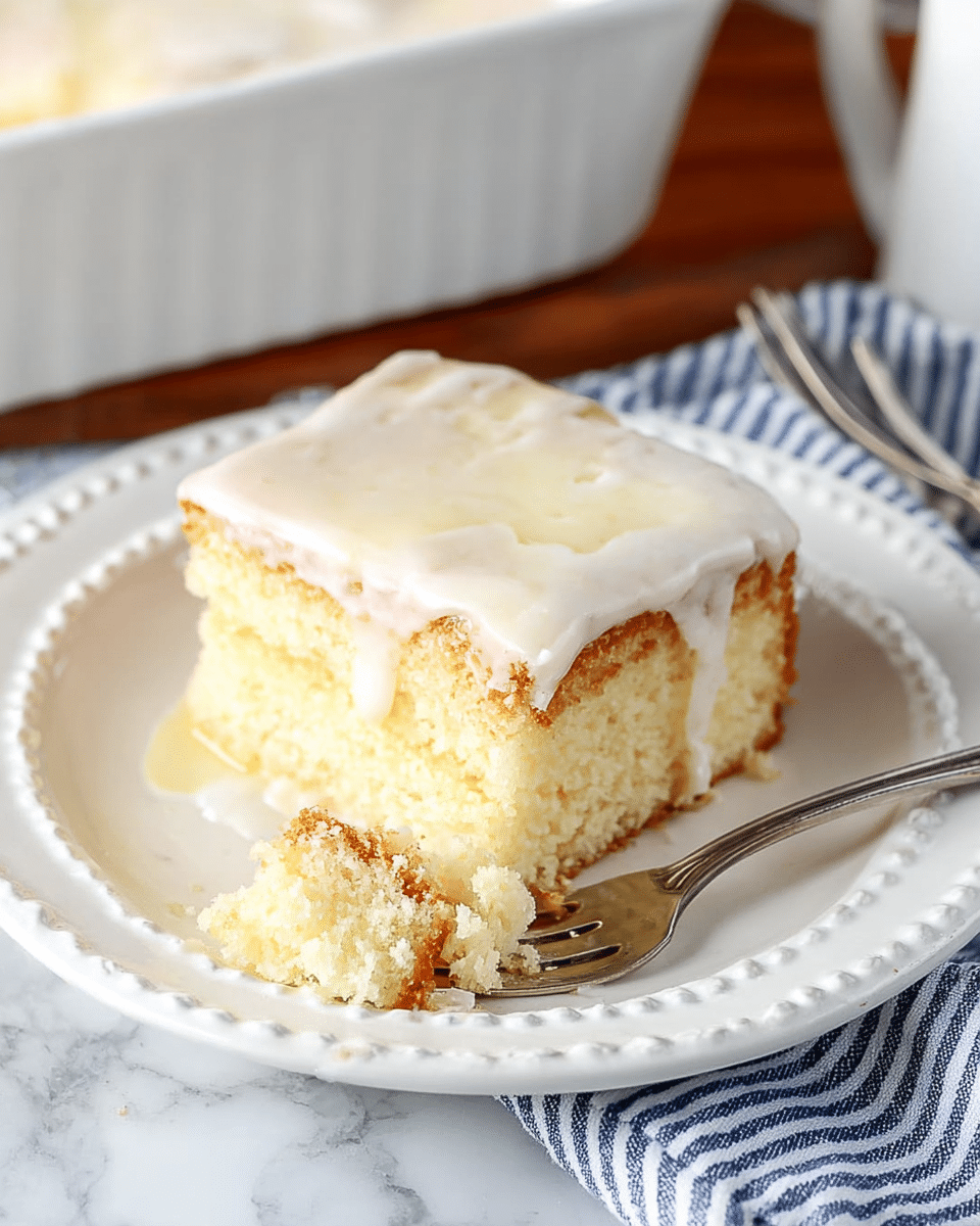 A close-up of a square piece of layered cake on a white plate with a beaded edge, placed on a white marbled surface. The cake has three visible layers: a light yellow bottom layer with a slightly browned edge, a thick middle layer that is creamy and pale, and a thin top layer covered in a glossy white icing that drips slightly down the sides. A bite-sized piece of the cake is on a silver fork resting on the plate, showing the soft and airy texture inside. The background includes a white casserole dish and a folded blue striped cloth with some silverware on it. Photo taken with an iphone --ar 4:5 --v 7