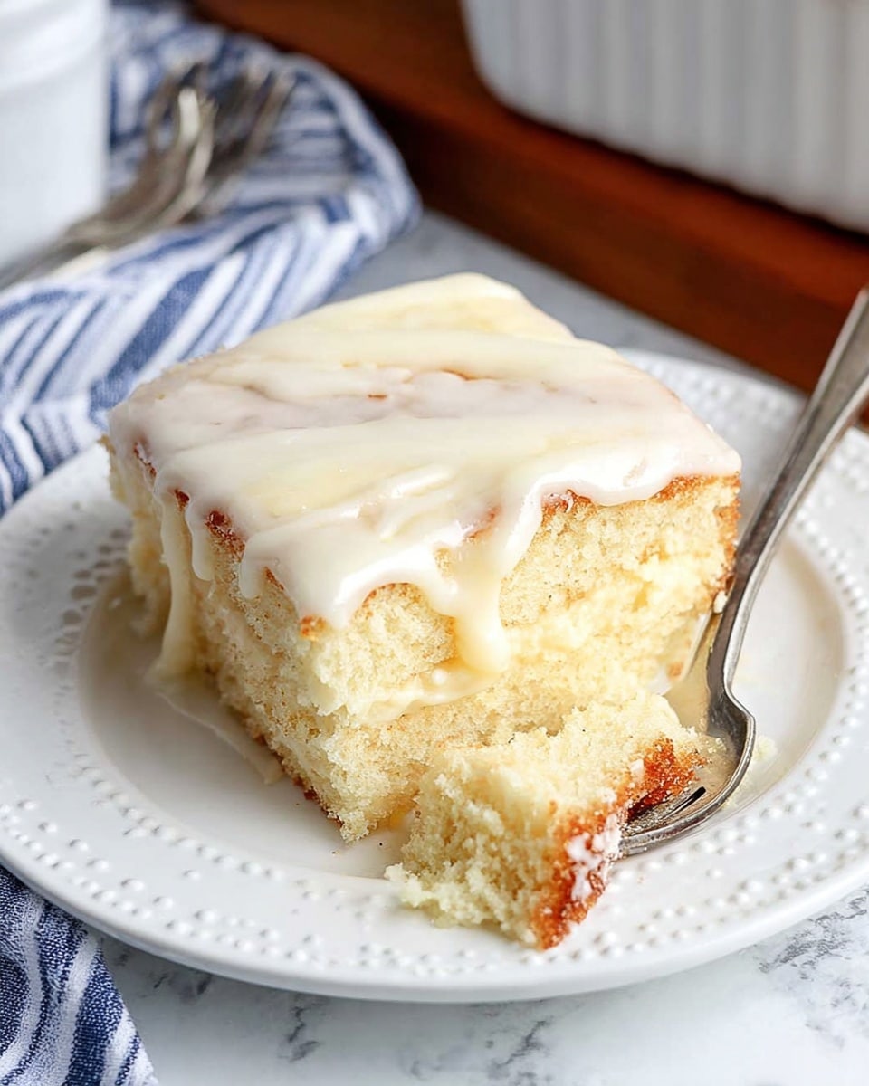 A single square piece of soft cake with three visible layers, each layer light cream in color and fluffy in texture; the top is covered with a smooth, white glaze that drips slightly down the sides. The cake sits on a white plate with a delicate dotted edge. A silver fork rests on the plate, holding a small bite of the cake with a brown crust bottom. The background shows a white marbled texture with a blue and white striped cloth and a hint of a white baking dish and silverware in the back. Photo taken with an iphone --ar 4:5 --v 7