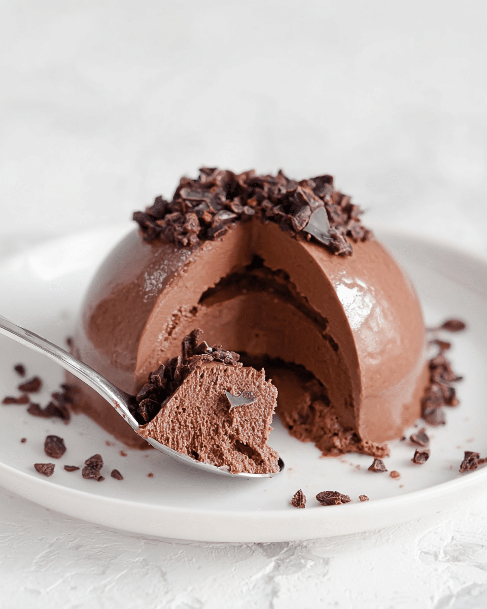 The image shows a smooth, rich chocolate mousse dessert shaped like a dome with a glossy surface, cut open to reveal its creamy texture inside. The mousse is dark brown and topped with small, rough pieces of cocoa nibs scattered on and around it, adding a crunchy texture contrast. A silver spoon slices through the mousse, lifting a piece that shines softly in the light. The dessert is placed on a clean white plate, set on a white marbled texture background, making the chocolate colors and textures stand out clearly. Photo taken with an iphone --ar 4:5 --v 7