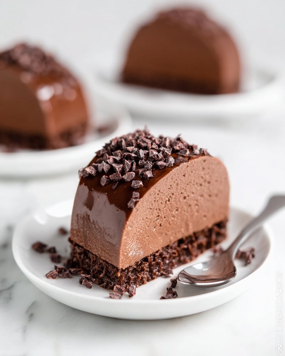 A smooth, rich chocolate dessert is shown with three curved layers stacked upright on a white plate; the layers are glossy and dark brown with a creamy texture, and the top and base are sprinkled with small, dark chocolate nibs. The plate sits on a white marbled texture, and a shiny spoon is placed nearby. In the blurred background, a similar chocolate dessert is visible on another white plate. Photo taken with an iphone --ar 4:5 --v 7