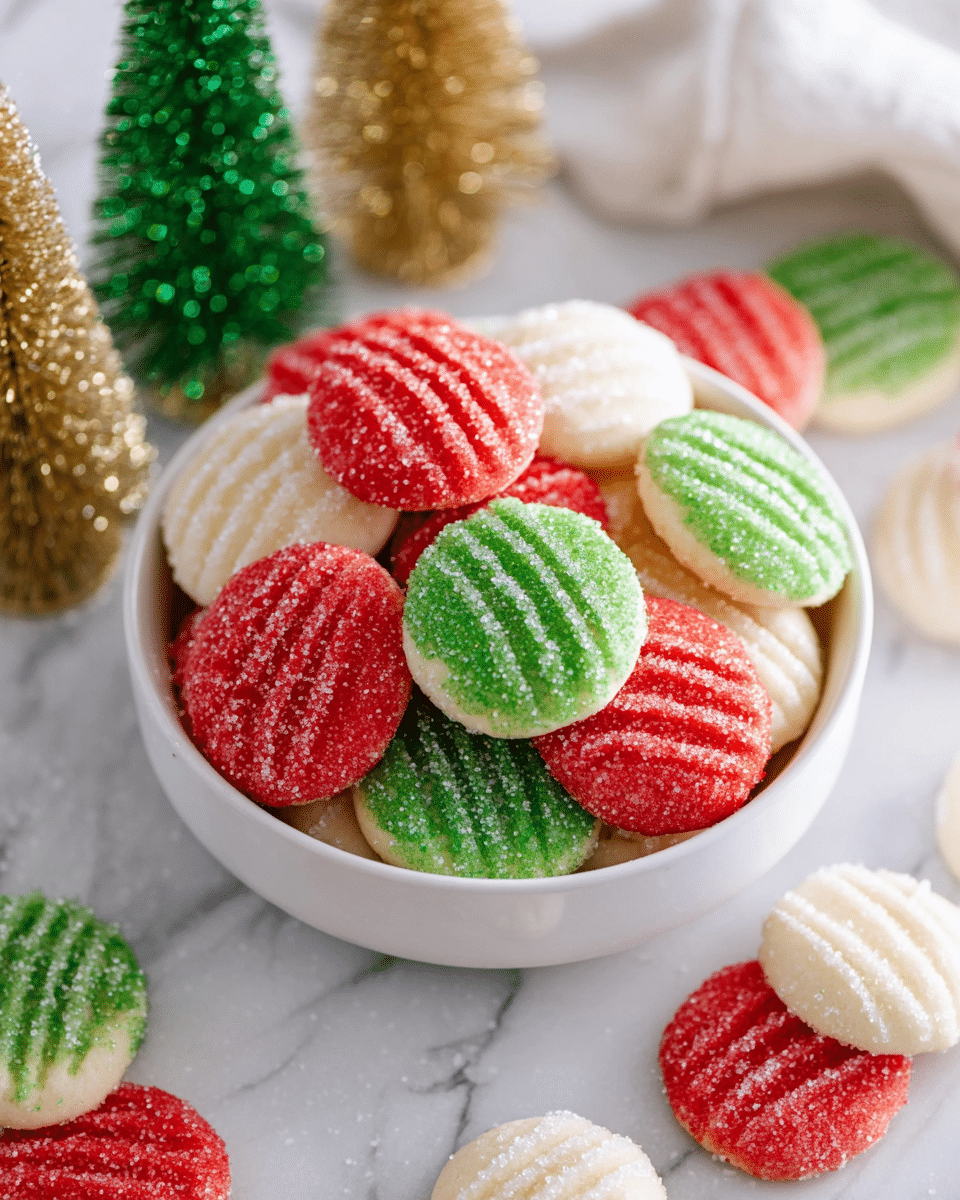 The image shows a white bowl filled with round sugar cookies in red, green, and white colors, each cookie having three shallow lines pressed across the top. The cookies have a light dusting of sugar crystals giving them a slightly sparkling texture. Around the bowl, there are a few cookies scattered on a white marbled surface. In the background, there are small decorative trees in green, gold, and white colors adding a festive touch. photo taken with an iphone --ar 4:5 --v 7