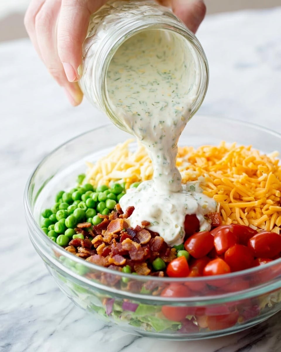 A woman's hand is holding a glass jar pouring thick, creamy white dressing with green herb specks over a clear glass bowl filled with colorful salad ingredients. The salad layers include bright green peas on the left, crispy brown bacon bits in the center, vibrant red grape tomatoes on the right, and shredded orange-yellow cheddar cheese on the far left, all arranged on a white marbled surface. photo taken with an iphone --ar 4:5 --v 7