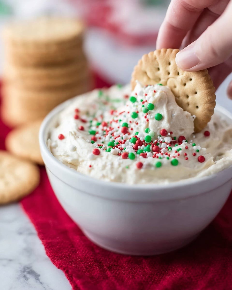A white bowl filled with white creamy dip topped with red, green, and white circular sprinkles. A woman’s hand is dipping a beige shortbread cookie with a detailed embossed pattern halfway into the dip, with the dipped part covered in the white creamy dip and colorful sprinkles. The bowl sits on a red cloth with blurred stacks of more cookies in the background, all set on a white marbled texture. Photo taken with an iphone --ar 4:5 --v 7