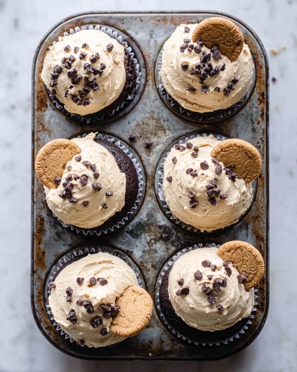A metal cupcake tray holds six chocolate cupcakes, each topped with a thick layer of light beige frosting mixed with small dark chocolate chips, giving it a slightly rough texture. Three of the cupcakes have a small round chocolate chip cookie placed on top, adding a golden brown color contrast. The cupcakes sit snugly in the tray, which shows signs of wear with some rust and dark spots. The background is a white marbled texture, creating a clean and bright look. Photo taken with an iphone --ar 4:5 --v 7