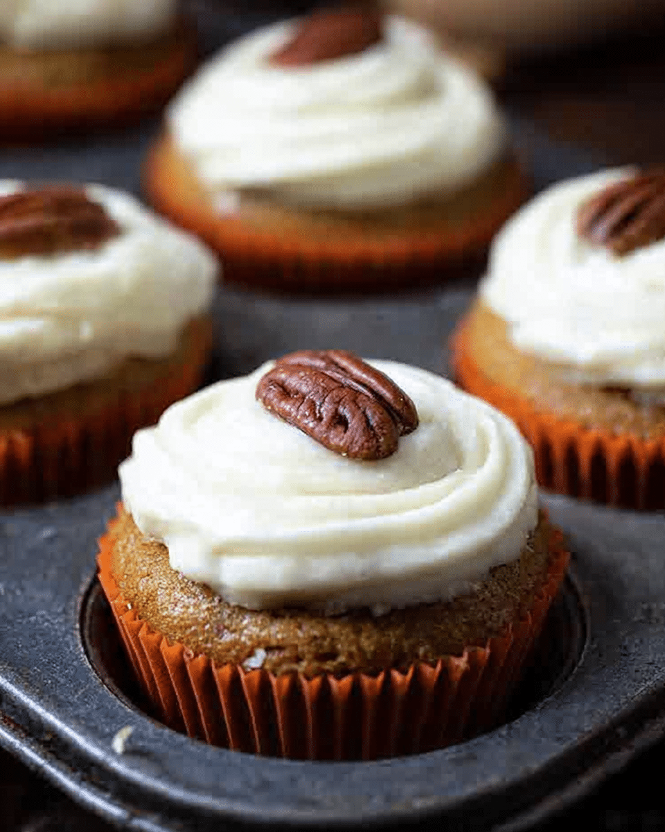 The image shows several cupcakes in a baking tray with a dark metal texture. Each cupcake has three visible layers: the bottom layer is a golden brown cake inside an orange paper liner, the middle layer is a thick, creamy white frosting swirled on top, and the top layer is a single whole brown pecan placed in the center of the frosting. The background and surface are not clearly visible, focusing attention on the cupcakes. Photo taken with an iphone --ar 4:5 --v 7