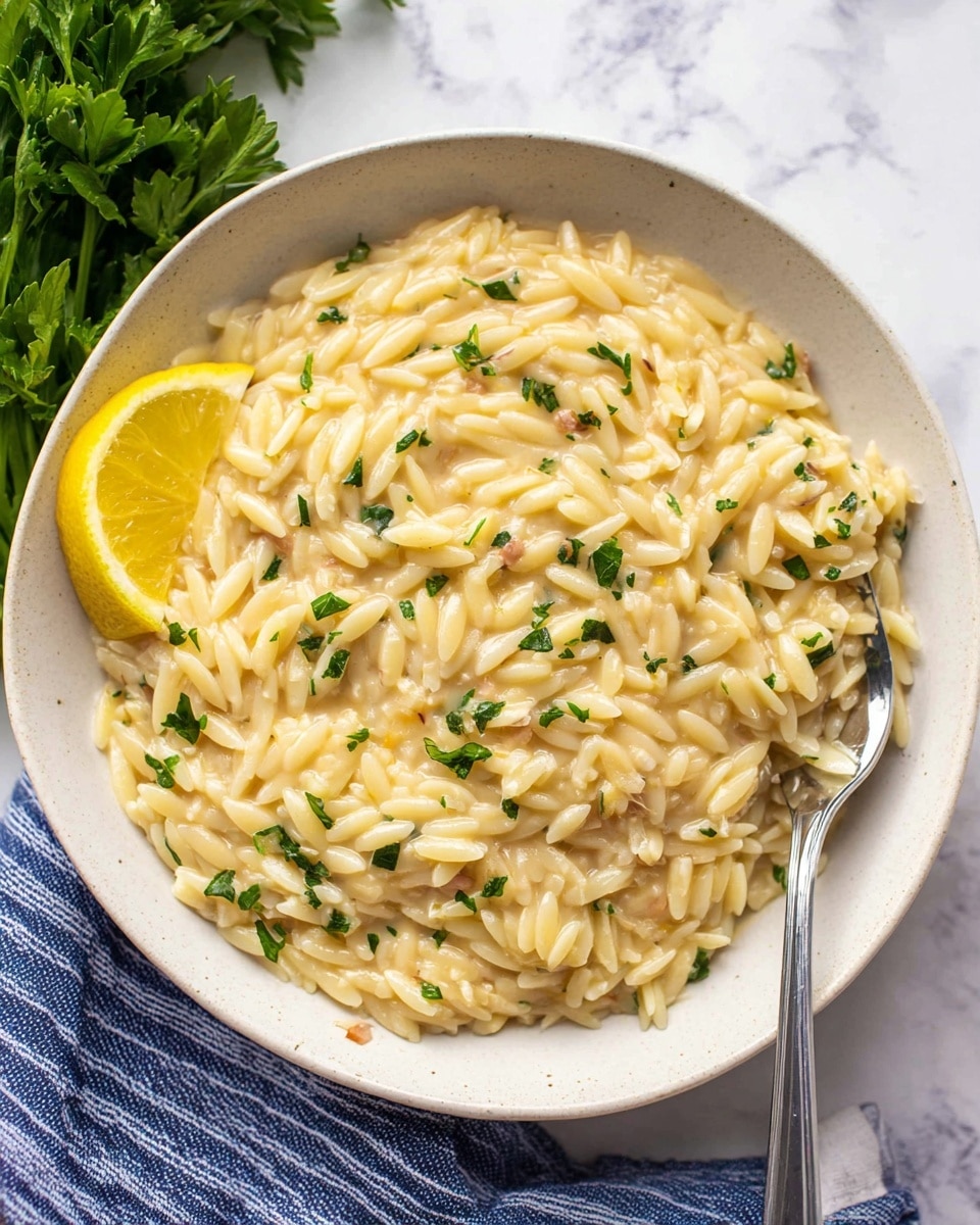 A white bowl filled with creamy orzo pasta, where the small rice-shaped pieces are coated in a smooth, light yellow sauce with a slightly glossy texture. The pasta is garnished with small green parsley leaves scattered evenly throughout. There is a slice of lemon placed on the left edge of the bowl, adding a vibrant yellow color. A silver fork is partially inserted into the orzo on the right side of the bowl, resting on top of the pasta. The bowl sits on a white marbled surface with a bunch of fresh parsley visible in the top left corner and a blue and white striped cloth partially underneath the bowl at the bottom left. photo taken with an iphone --ar 4:5 --v 7