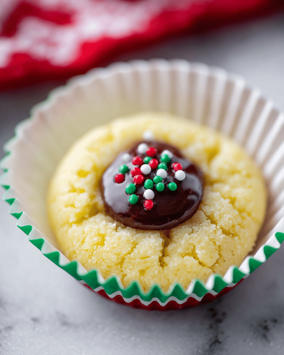 A soft, round yellow cookie sits in a white cup liner with green and red edges, showing a slightly cracked texture on top. It has a shiny dark brown chocolate dollop in the middle, decorated with tiny round sprinkles in red, green, and white. The cookie is placed on a white marbled surface, with a blurred red and white cloth in the background. photo taken with an iphone --ar 4:5 --v 7