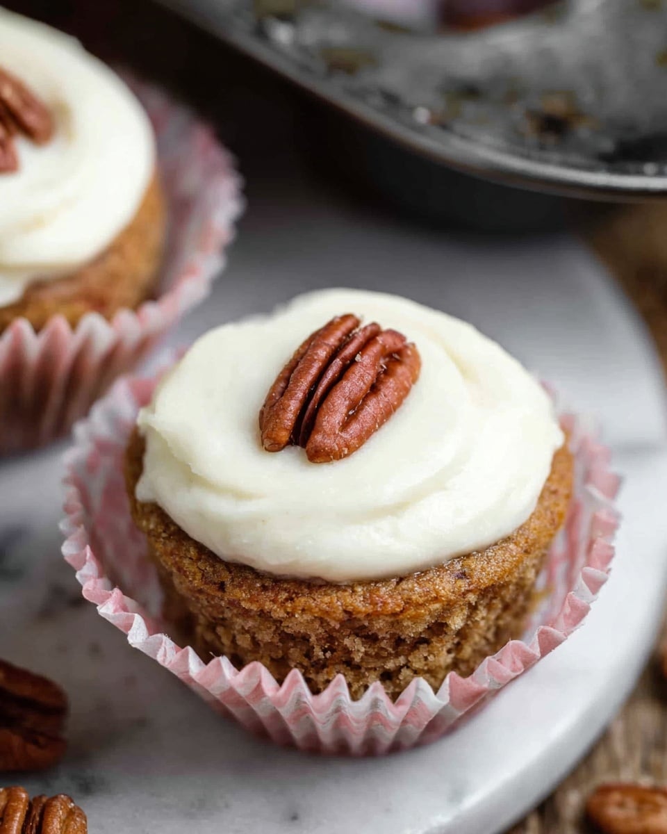 A close-up view of a single cupcake with two main layers, starting with a light brown, textured base that looks moist and slightly crumbly, held inside a pink and white striped cupcake liner. On top of the base, there is a smooth, thick layer of white frosting with a creamy texture, dolloped in a circular shape. At the center of the frosting, there is a pecan half resting flat, adding a crunchy detail and a deeper brown color contrast. The cupcake is on a white marbled surface, with parts of a metal cupcake tray and more pecans gently blurred in the background. photo taken with an iphone --ar 4:5 --v 7
