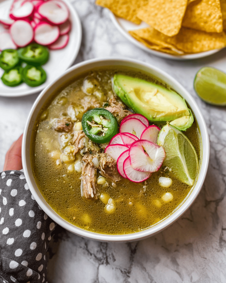 A white bowl filled with green broth soup that has visible chunks of tender meat, white corn kernels, and a slightly oily surface. On top, there are thinly sliced bright pink-edged radish rounds arranged in a small pile, a slice of green jalapeno pepper with seeds showing, a pale green wedge of lime on the right side, and a thick light green slice of avocado resting on the left rim of the bowl. The bowl is set on a white marbled texture with scattered yellow corn tortilla chips in the background and a white plate holding extra radish and jalapeno slices. A woman's hand with a polka dot fabric is partially visible in the bottom left corner. Photo taken with an iphone --ar 4:5 --v 7