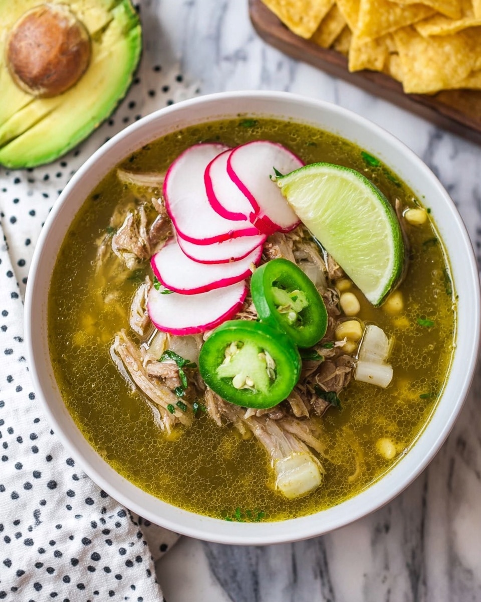 A white bowl filled with green broth holding shredded meat, light beige corn pieces, and topped with thinly sliced pink and white radishes, a few green jalapeño slices, a bright green lime wedge, and a quarter of a ripe avocado with smooth light green flesh, all placed on a white marbled surface with a white cloth featuring black polka dots nearby and some yellow tortilla chips partially visible in the background; photo taken with an iphone --ar 4:5 --v 7