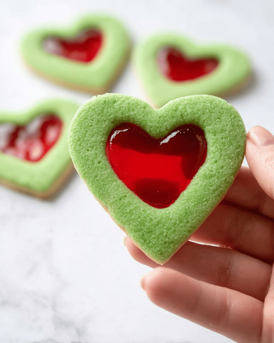 A close-up of a green heart-shaped cookie with a smaller red heart-shaped cutout filled with a shiny, translucent red layer in the center. The cookie has a soft, slightly textured surface and is held by a woman's hand at the bottom left of the image. In the background, there are three more similar green heart cookies with red heart centers lying flat on a white marbled surface, slightly blurred. The overall image is bright and clear with a simple, clean setting. photo taken with an iphone --ar 4:5 --v 7