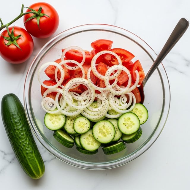 A clear glass bowl filled with a fresh salad showing three visible layers: the bottom layer is light green cucumber slices with soft texture, the middle layer is bright red tomato wedges with a juicy texture, and the top layer is thin white onion rings spread evenly. The vegetables have black pepper sprinkled over them. A metal spoon rests inside the bowl on the right side. The bowl sits on a white marbled surface with a whole green cucumber and two red tomatoes with green stems placed on the left side. Photo taken with an iphone --ar 4:5 --v 7