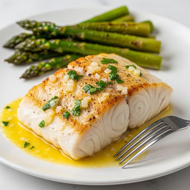 A thick, white piece of fish cooked with a light golden brown crust on top, covered in a shiny, yellow butter sauce with small bits of green herbs and white onions scattered on the surface, resting on a white plate. Behind the fish, a small pile of cooked asparagus with a slightly charred green texture is visible. A silver fork with thin prongs touches the bottom right corner of the fish. The entire setting is on a white marbled texture. photo taken with an iphone --ar 4:5 --v 7