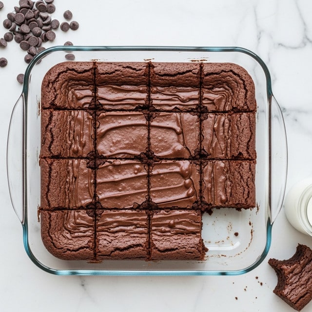 A clear glass square baking dish contains 12 cut chocolate brownies arranged in a 4 by 3 grid, with one piece missing from the lower right corner. The brownies have a smooth, dark brown top layer with slight wrinkles and a dense, moist texture inside. The dish is placed on a white marbled surface. Near the top left corner, there is a small pile of dark chocolate chips. A small glass of milk and a piece of brownie with a bite taken out of it are visible at the bottom right. The photo taken with an iphone --ar 4:5 --v 7