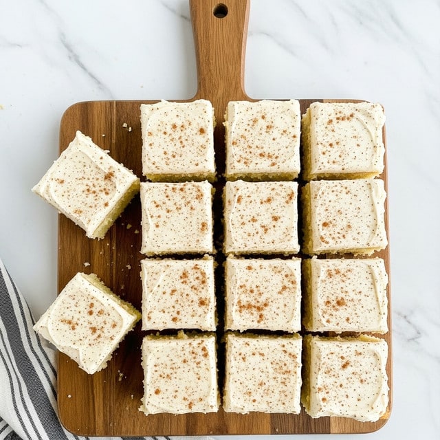 There are sixteen square cake pieces arranged in a 4 by 4 grid on a wooden cutting board with a handle at the top center. Each piece has a thick, creamy white frosting layer on top, sprinkled evenly with small brown specks, likely cinnamon or nutmeg. The cake underneath looks soft and light yellow, slightly textured and porous. Two pieces on the left and one on the bottom right are slightly separated from the others. The board is placed on a white marbled surface, and a black and white striped cloth is partially visible at the bottom left corner. Photo taken with an iphone --ar 4:5 --v 7