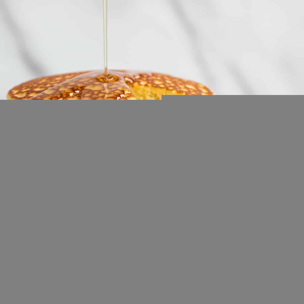 A stack of seven light golden, porous pancakes with a soft, spongy texture sits on a white plate, with a slice cut out to show the layers. Each pancake layer is thin and even, revealing the airy texture inside. A light syrup is poured over the top and gently drips down the sides, pooling slightly at the base on the plate. The background shows a soft, white marbled texture. photo taken with an iphone --ar 4:5 --v 7