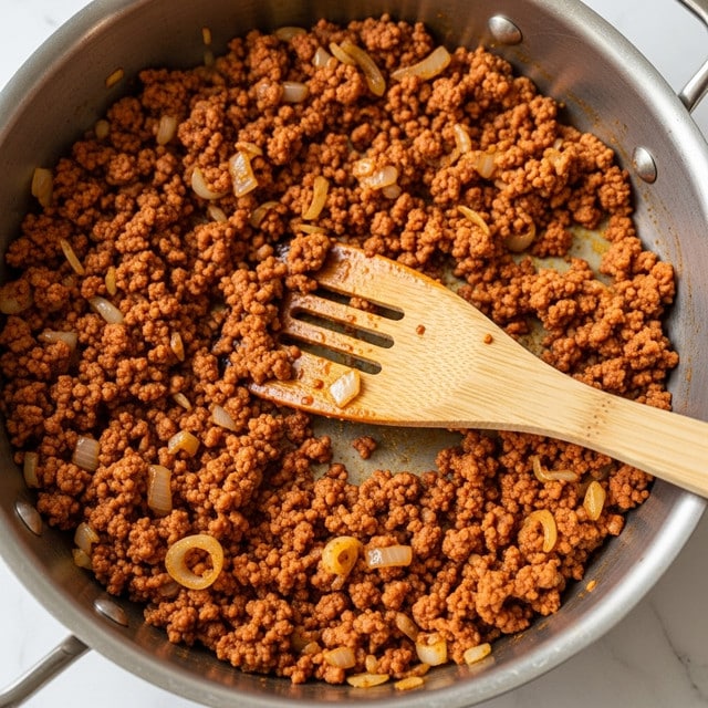 The image shows a close-up of a large metal pan filled with finely ground cooked meat mixed with small pieces of sautéed onions. The meat has a crumbly texture with a reddish-brown color, and the onions are soft with a light golden shade, all coated with a slightly oily seasoning. A wooden spatula with a natural wood grain texture rests inside the pan, partly covered with the same meat mixture. The background has a white marbled texture. photo taken with an iphone --ar 4:5 --v 7