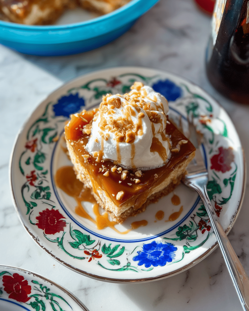 A square slice of layered dessert sits on a white plate decorated with blue and red flowers and green leaves on the edges; the dessert has a golden-brown crust base visible underneath a thick rich caramel layer with a glossy texture, topped with a dollop of white whipped cream that is smooth and fluffy, sprinkled with small chopped nuts and drizzled with a light caramel sauce. A silver fork rests on the right side of the plate. The plate is placed on a white marbled surface with parts of a dark bottle and a blue bowl with similar dessert visible at the top of the image. Photo taken with an iphone --ar 4:5 --v 7