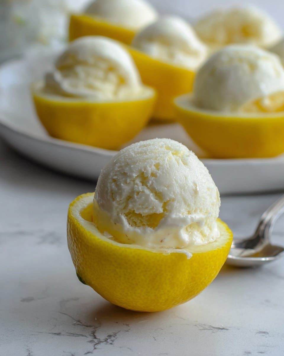 This image shows a close-up of a small yellow lemon half serving as a natural bowl, filled with a creamy white scoop of ice cream that has a smooth yet slightly textured surface. The lemon bowl sits on a white marbled surface with a slightly shiny and fresh look. In the background, there are several more lemon halves also filled with white ice cream, arranged on a white plate which adds depth but is softly blurred, keeping focus on the front lemon half. A silver spoon is partly visible in the background, lying on the surface behind the lemon bowl. photo taken with an iphone --ar 4:5 --v 7