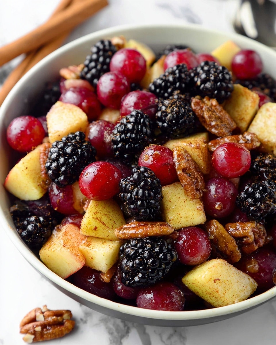 A close-up of a white bowl filled with a colorful fruit salad showing multiple layers of different fruits and nuts. The bottom layer has shiny blackberries, their round, bumpy deep black texture visible. On top are smooth, round red grapes, glistening with moisture. Large pieces of apple chunks are scattered throughout, coated with a sprinkle of brown spices. Among the fruits are crunchy, golden brown pecan halves adding texture and contrast. The bowl sits on a white marbled surface, with two cinnamon sticks placed nearby. Photo taken with an iphone --ar 4:5 --v 7