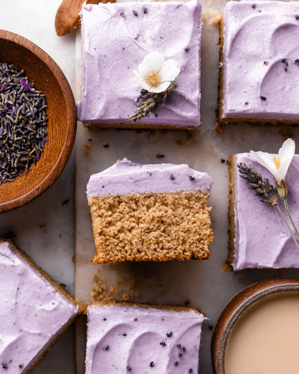 A close-up view of a soft, spongy square cake slice showing two clear layers: the bottom layer is a light brown cake with a slightly crumbly texture, speckled with tiny darker bits, while the top layer is thick, smooth, and pale purple frosting spread evenly across the cake. On top of the frosting rests a small white flower and a sprig of light purple lavender, adding a delicate touch. In the background, similar cake slices are visible, all with the same two-layer structure, set on a light wooden surface beside a white marbled texture. A wooden bowl with dried lavender sits to the side, enhancing the natural and fresh feeling of the scene. photo taken with an iphone --ar 4:5 --v 7