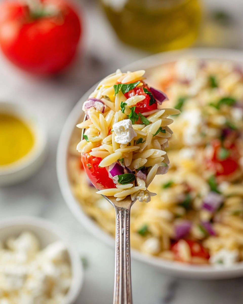 This image shows a close-up of a spoon holding a scoop of orzo pasta salad. The salad has three main layers: pale yellow orzo pasta as the base, mixed with small pieces of red tomato and purple onion scattered throughout, and white crumbly feta cheese on top, all garnished with small green parsley leaves. In the blurred background, a white bowl with the rest of the salad can be seen on a white marbled surface, along with a small white bowl containing feta cheese and a bottle of olive oil. Photo taken with an iphone --ar 4:5 --v 7