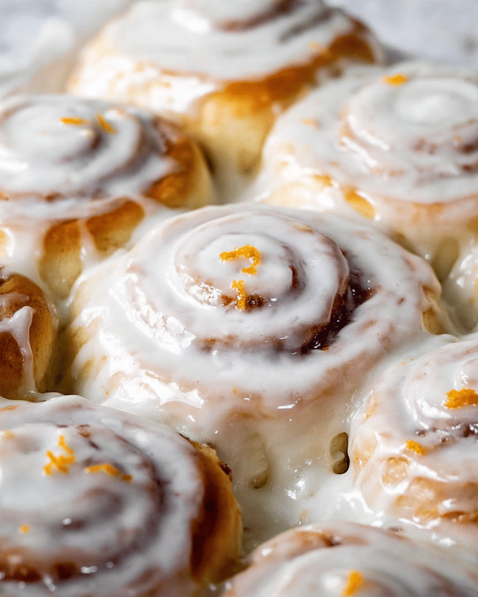 The image shows a close-up of soft cinnamon rolls covered with a thick white glaze. There are about nine cinnamon rolls arranged closely together in one layer, with their spiral shapes visible under the glaze. The glaze has a smooth, shiny texture and covers the rolls unevenly, dripping down the sides. Small bits of orange zest are sprinkled on top, adding flashes of bright orange color against the white glaze. The whole scene is set on a white marbled surface, giving a clean and fresh look. photo taken with an iphone --ar 4:5 --v 7
