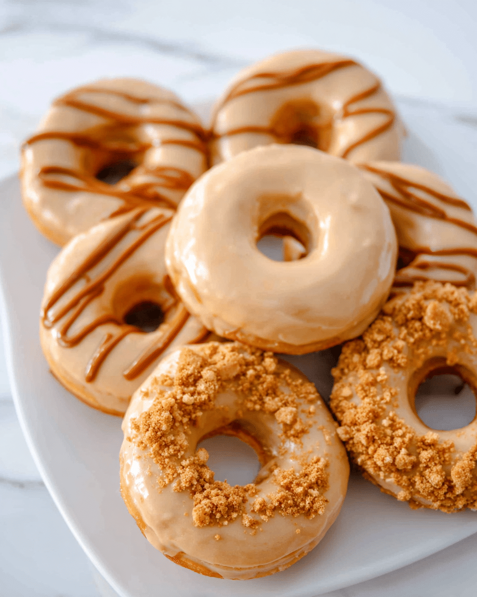 The image shows six donuts arranged in two rows on a white rectangular plate set on a white marbled surface. The donuts have a light golden-brown base with a smooth, creamy tan glaze. Three donuts are topped with crumbly golden brown bits scattered unevenly over the glaze, giving a rough, textured look. Two donuts have a similar tan glaze but are decorated with diagonal drizzles of a darker caramel-colored icing and some crumbly bits on top. One donut in the top center has a plain tan glaze without any toppings, looking smooth and shiny. The donuts' soft, cozy texture contrasts with the crunchy toppings and smooth icing. photo taken with an iphone --ar 4:5 --v 7