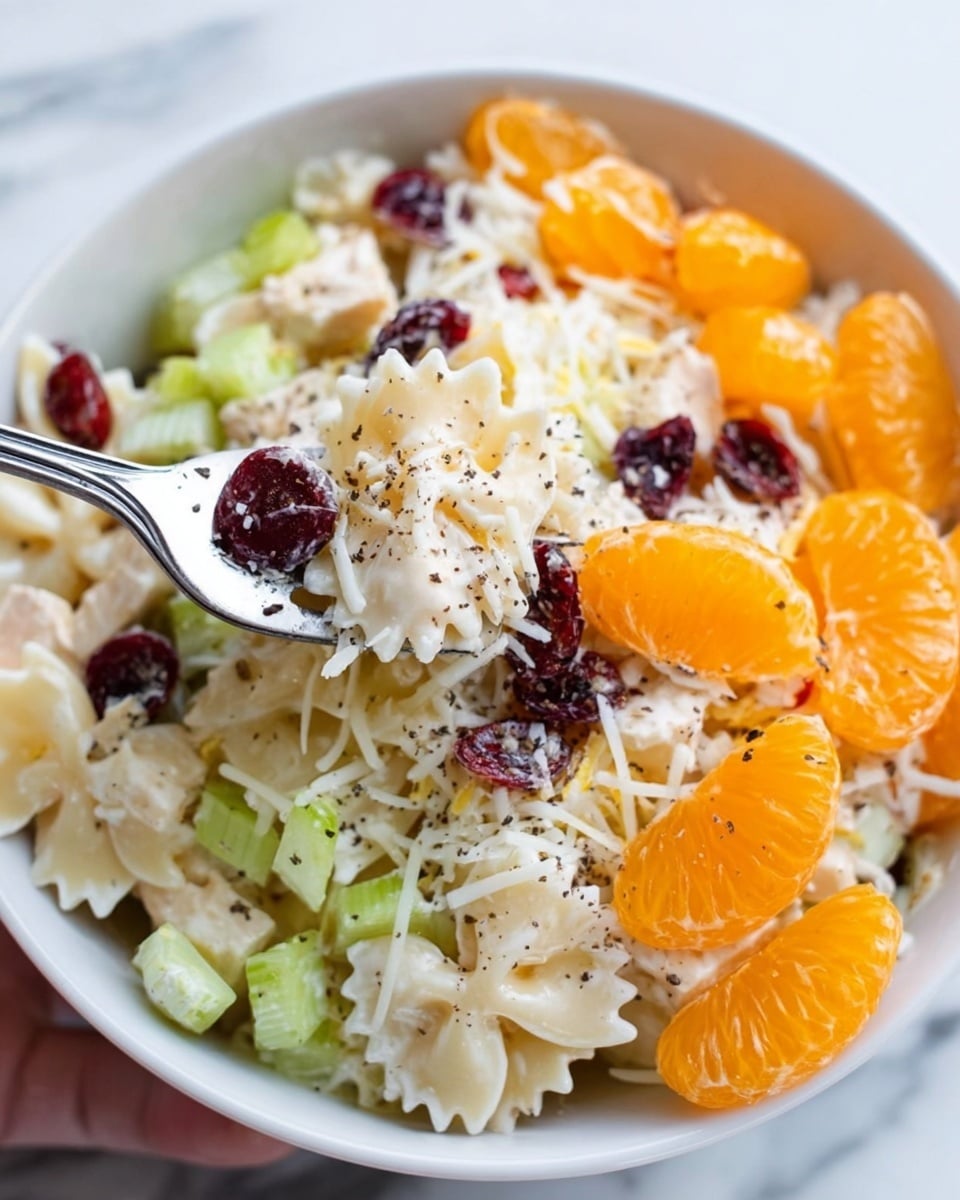A close-up of a white bowl filled with a colorful fruit pasta salad on a white marbled texture. The salad has 4 visible layers: the base layer is creamy white bow-tie pasta mixed with shredded white cheese; the second layer includes small chunks of pale green celery pieces scattered evenly; the third layer features bright orange mandarin segments adding a juicy texture; and the top layer is sprinkled with dark red dried cranberries and freshly ground black pepper, giving contrast and texture. A silver fork held by a woman's hand is digging into the salad from the top left side. photo taken with an iphone --ar 4:5 --v 7