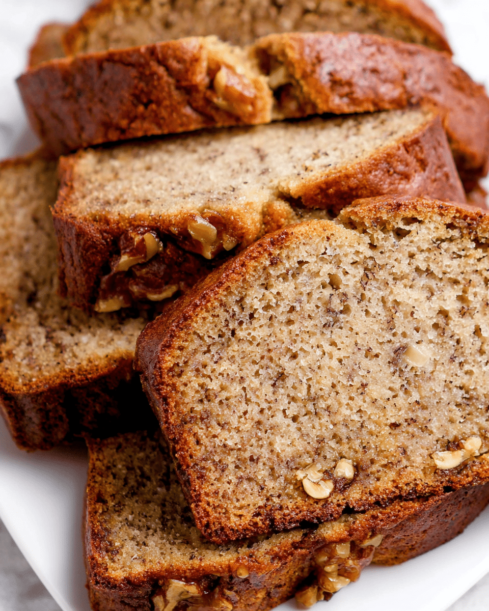 A close-up image shows several slices of moist banana bread stacked closely on a white plate, placed on a white marbled surface. The banana bread has a golden brown crust with a soft, light brown inside speckled with small chunks of walnuts, giving it a textured look. The slices vary slightly in thickness, and the bread’s crumb appears dense yet tender, with some cracks on the surface highlighting its homemade nature. photo taken with an iphone --ar 4:5 --v 7