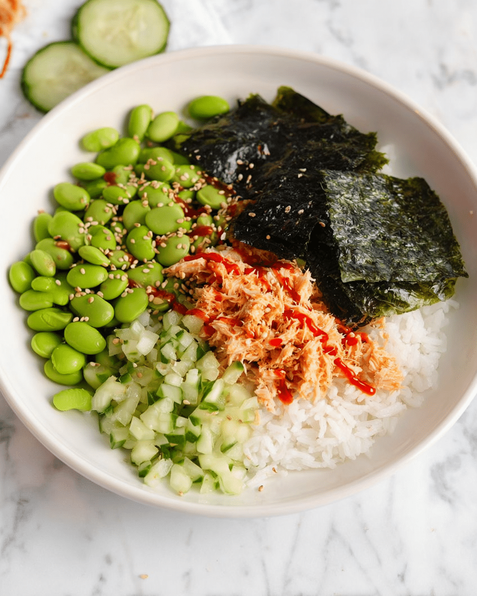 A white bowl on a white marbled surface holds a colorful layered dish starting with a base of white rice. On top of the rice, there are four distinct sections: bright green edamame beans with sesame seeds sprinkled on them, dark green seaweed sheets lightly sprinkled with sesame seeds placed neatly on one side, finely chopped light green cucumber pieces, and a creamy, shredded orange mixture that looks like tuna with small spots of red sauce drizzled over the layers. The arrangement is neat and each ingredient forms a clear section against the rice base. photo taken with an iphone --ar 4:5 --v 7