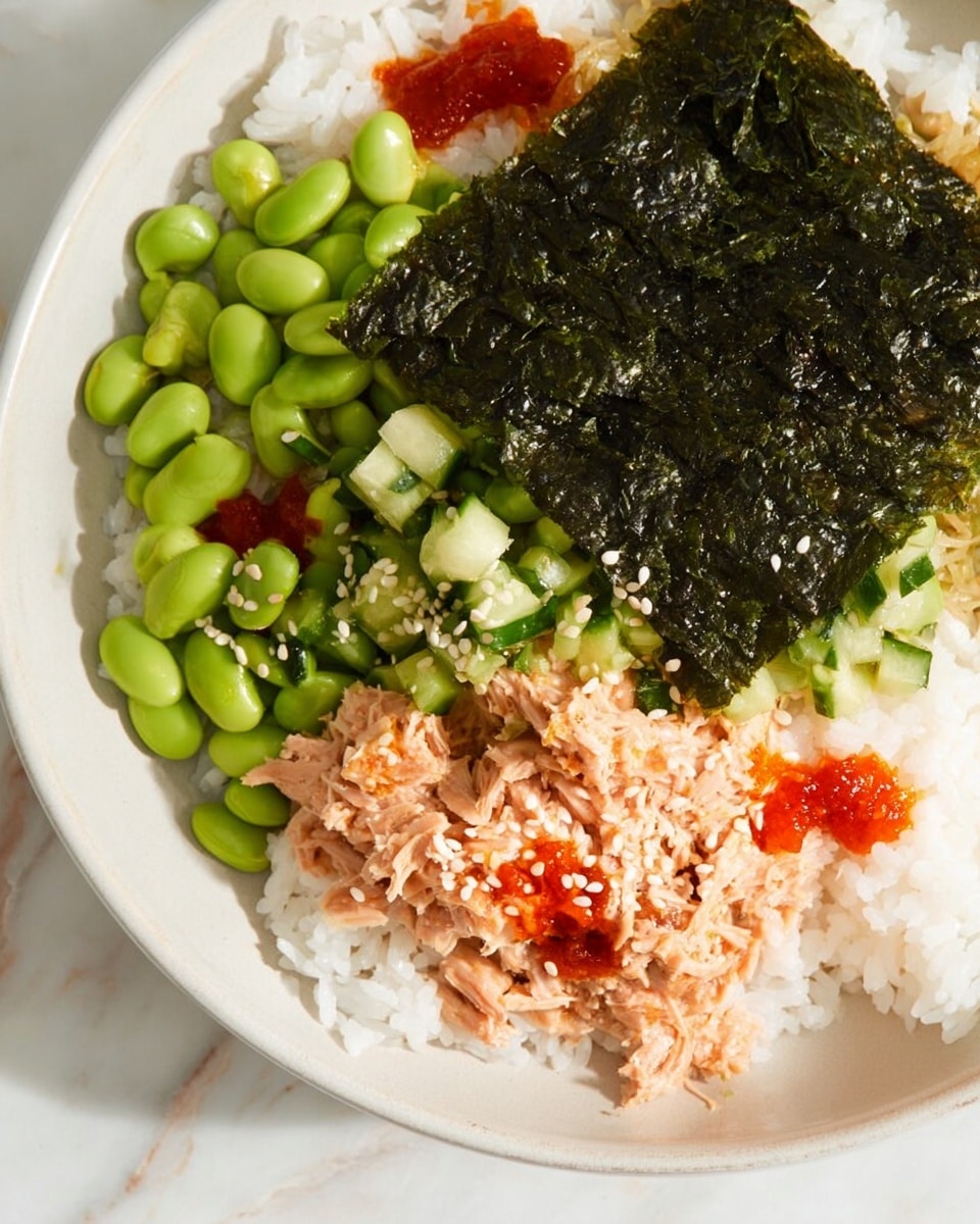 The dish is presented in a white bowl resting on a white marbled surface. The bottom layer consists of white rice, spread evenly across the bowl. On one side, there is a portion of light pink tuna salad with a soft, flaky texture, dotted with small red sauce spots. Next to that, bright green edamame beans sit in a cluster, glistening slightly with sesame seeds sprinkled on them. Adjacent to the edamame is a heap of chopped green cucumber pieces. At the top, a square sheet of dark green seaweed with a crinkly texture is placed, sprinkled with white sesame seeds. Small dots of red sauce are scattered over the dish, adding contrast and color. Photo taken with an iphone --ar 4:5 --v 7